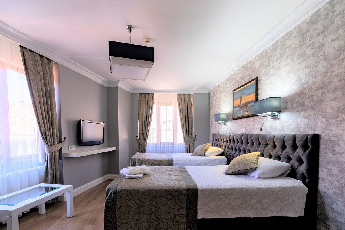 Una camera d'albergo con due letti, una televisione su un ripiano, un quadro al muro e una panca vicino alla finestra.