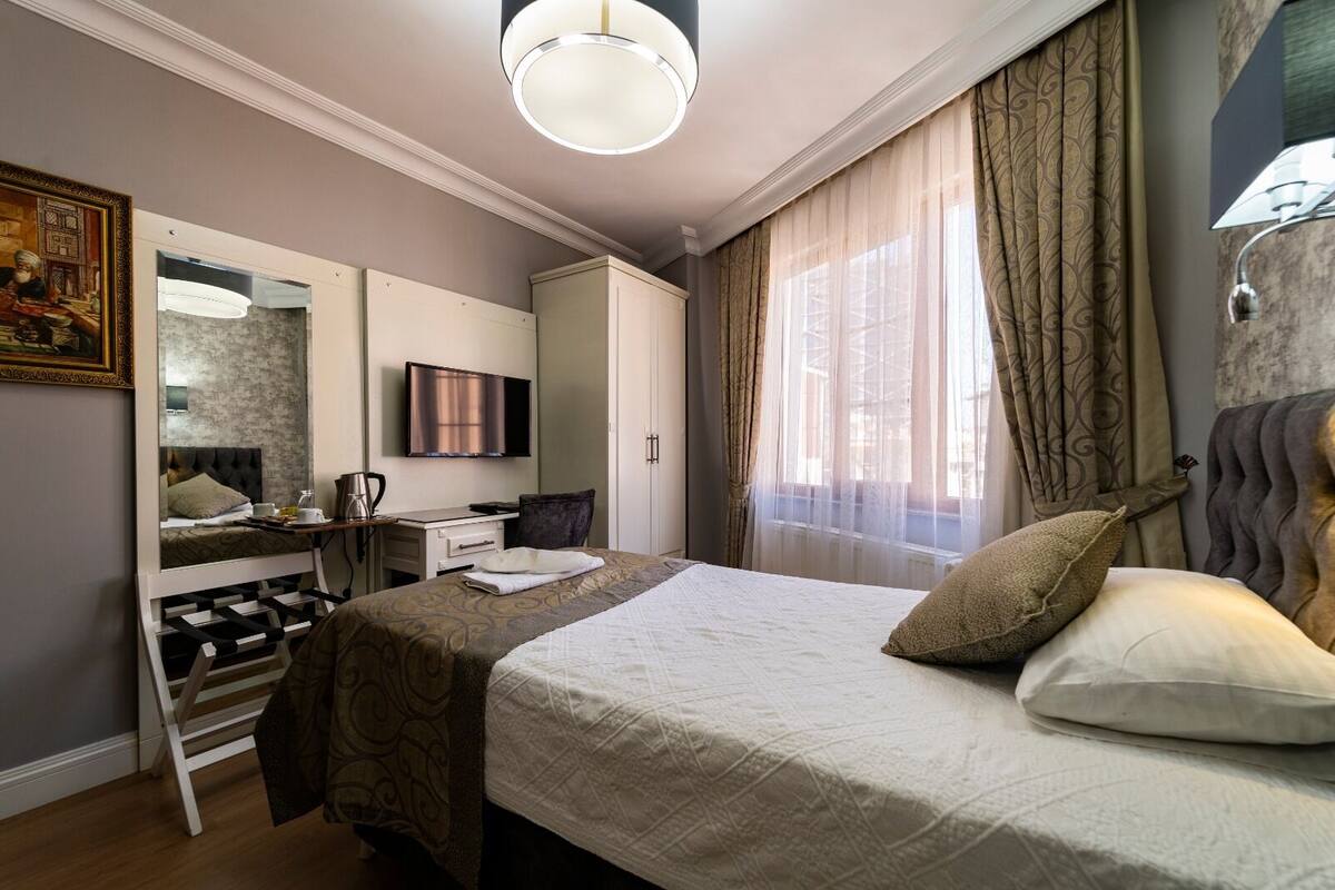 A hotel room with a bed, a desk, a television, and a large window with curtains.