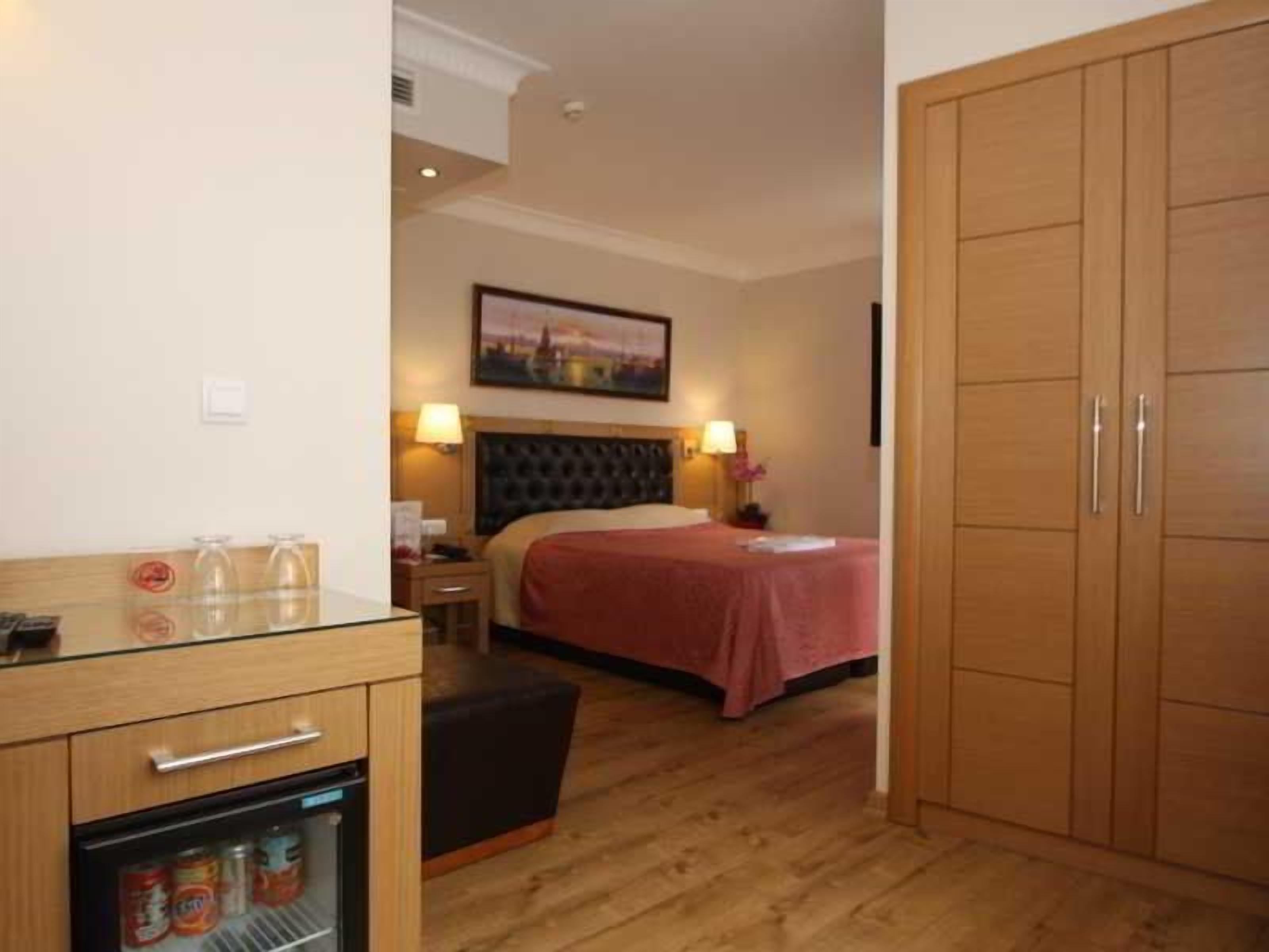 Quarto de hotel com cama, abajures, guarda-roupa de madeira, mesa pequena com tampo de vidro e mini-geladeira.