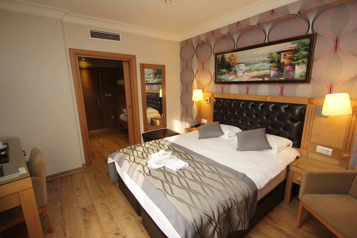 A hotel room with a large bed, two bedside tables, a desk, a chair, and a painting on the wall.