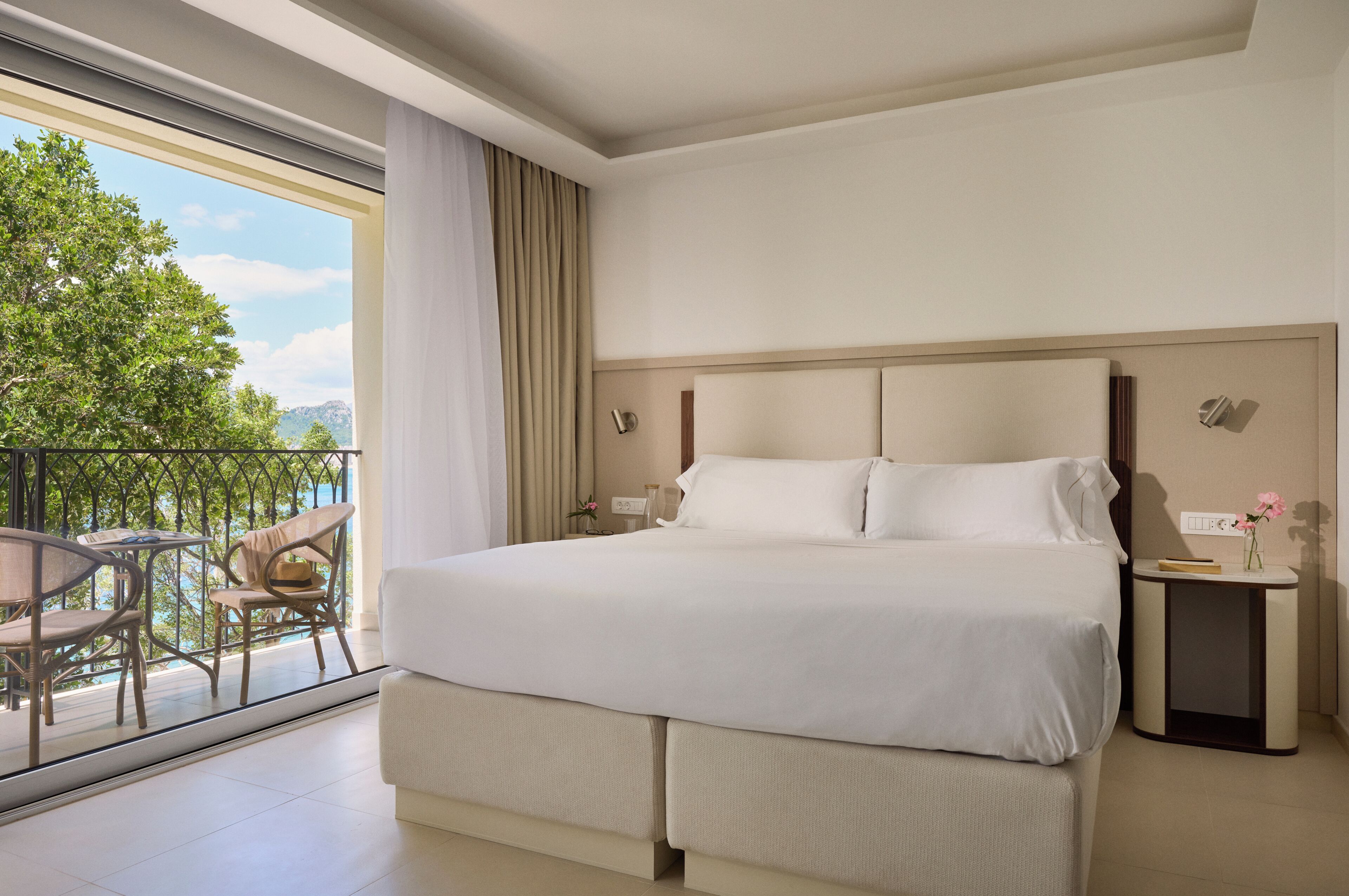 A hotel room with a large bed, a bedside table, a chair, a small table with a vase, and a balcony with chairs and a view of greenery.