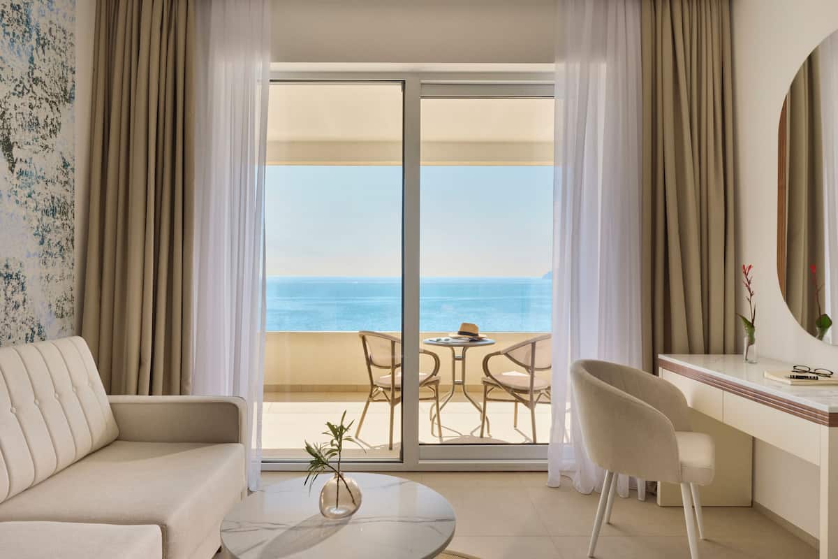 A hotel room with a sofa, a desk, a chair, a small table, and a view of the sea through a glass door.