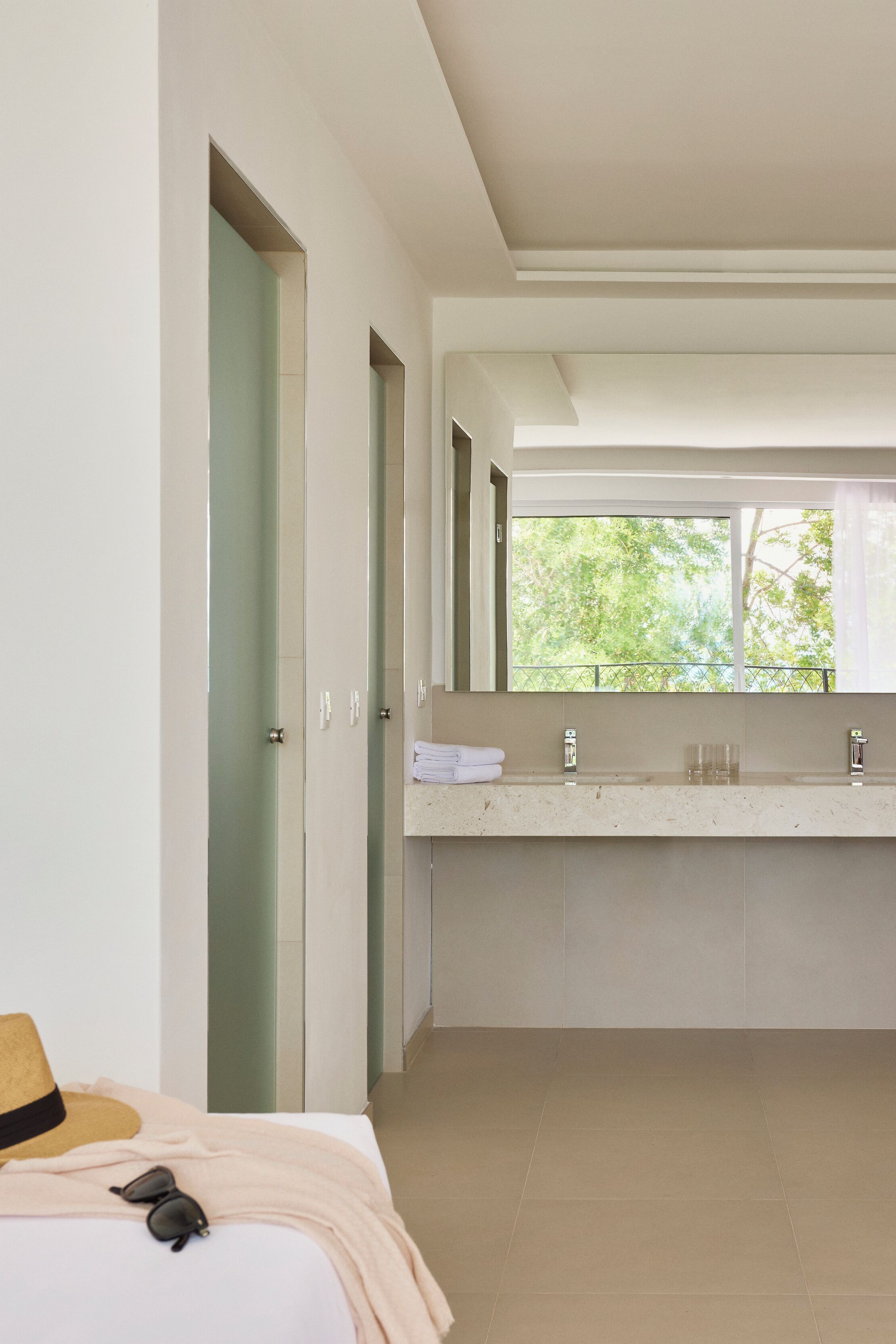 A modern bathroom with a large mirror, a window with a view of trees, and a bed with a hat and sunglasses on it.