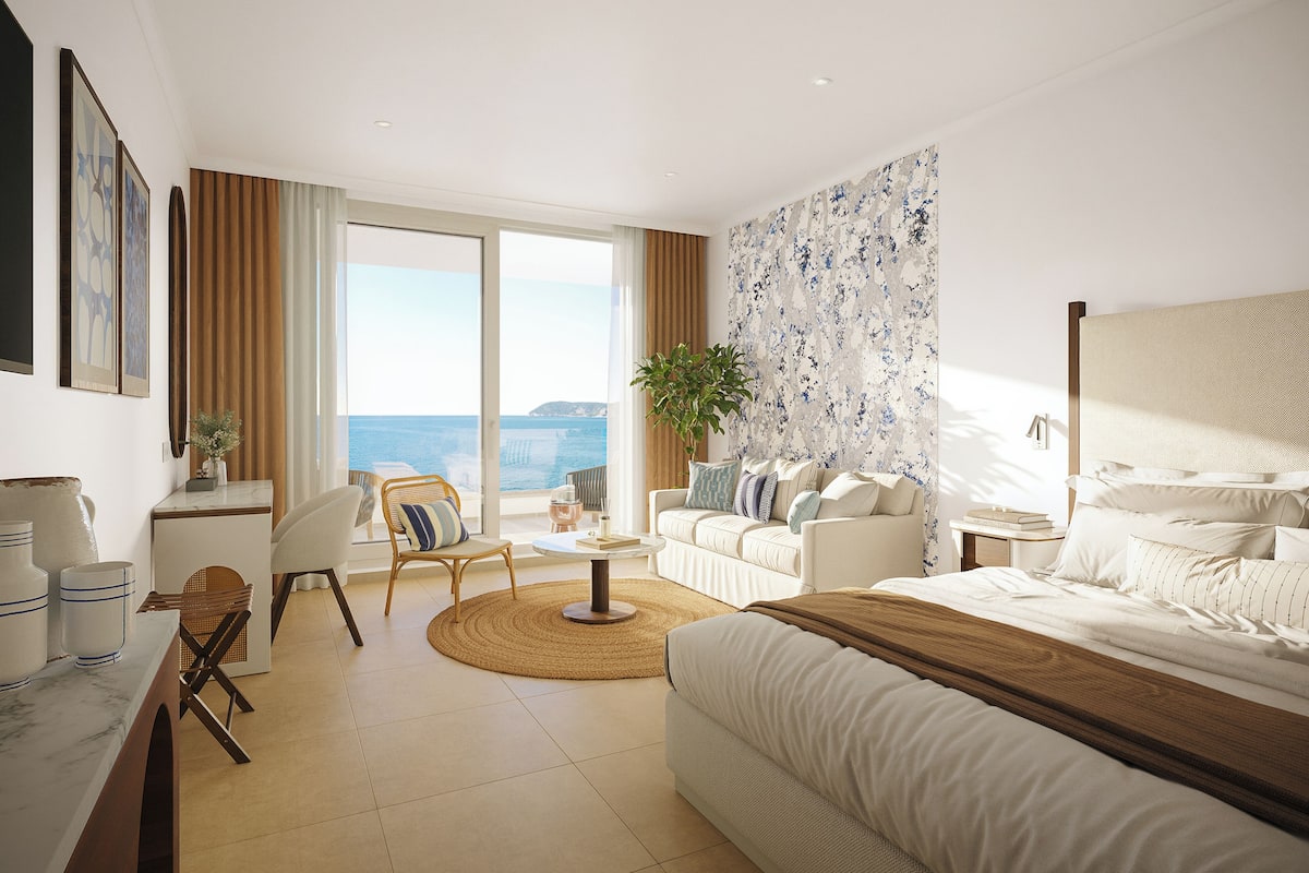 A modern hotel room with a large bed, a sofa, a desk, and a balcony with a view of the sea.