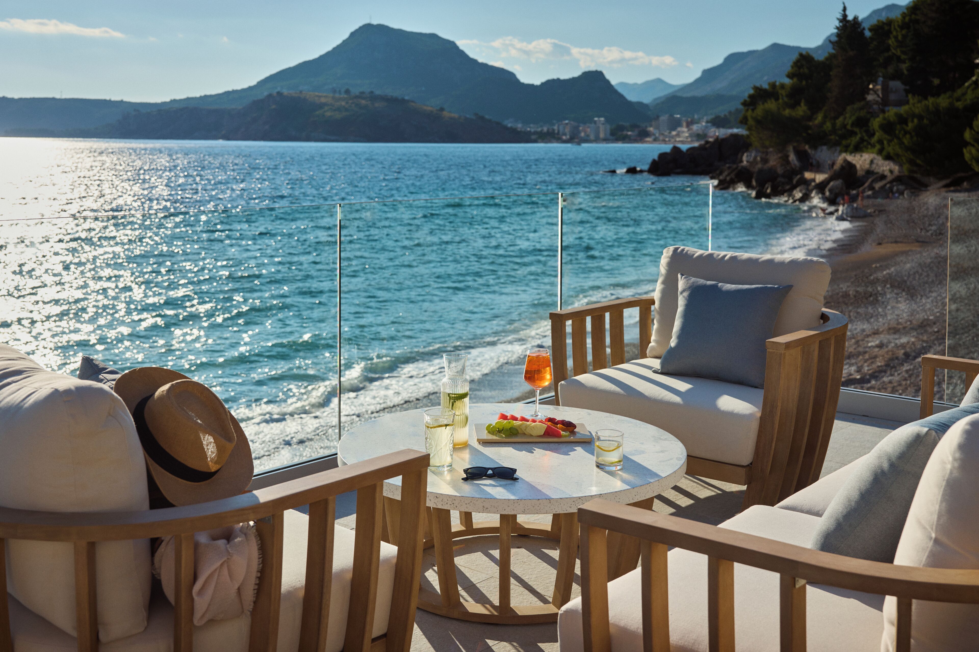 A seaside terrace with a round table, two chairs, and a view of the sea and mountains.