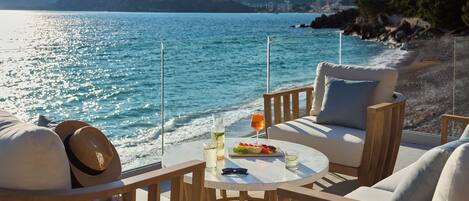 A seaside terrace with a round table, two chairs, and a view of the sea and mountains.