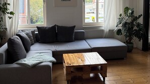 Ein modernes Wohnzimmer mit einem grauen Ecksofa, einem Holztisch und einem großen Fenster mit Vorhängen.