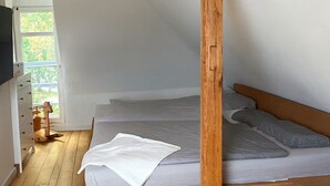 Ein kleines Schlafsofa mit Holzrahmen, grauem Lattenrost und Kissen, Holzboden und ein Fenster mit Blick auf Grünflächen.
