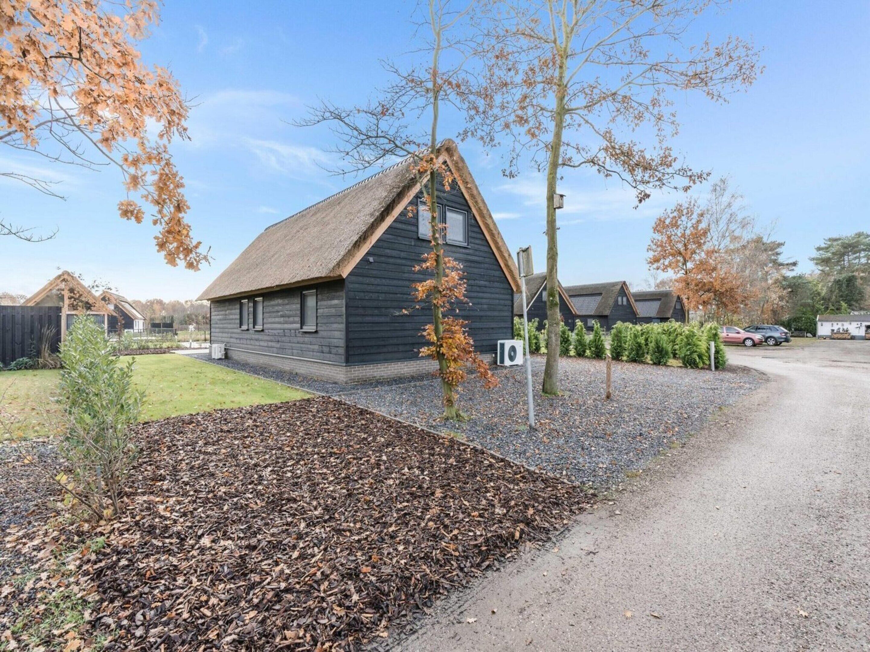 Un bâtiment moderne à l’extérieur sombre, surmonté d’un toit de chaume, entouré d’une allée de gravier et d’un sol recouvert de feuilles.