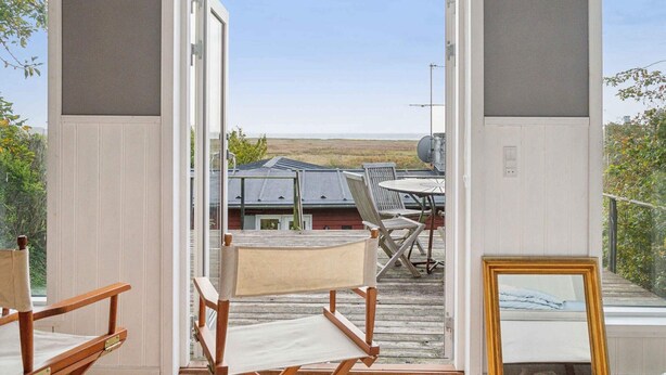 Une porte ouverte donne sur une terrasse où se trouvent une table et des chaises, et d’où l’on peut admirer un paysage avec un bâtiment visible au loin.