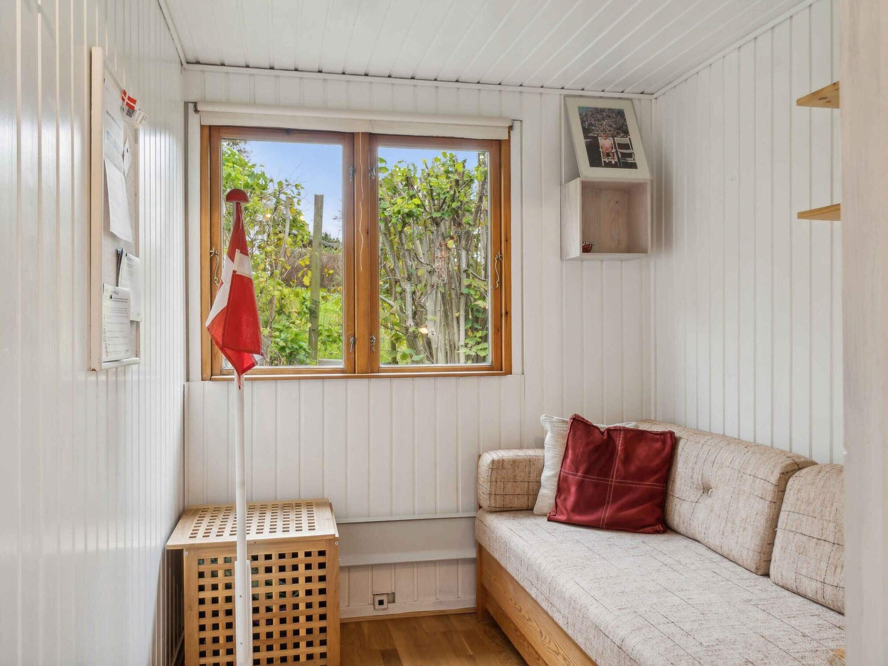 A room with a wooden floor, a white sofa with a red cushion, a window with a view of greenery, and a small wooden shelf.