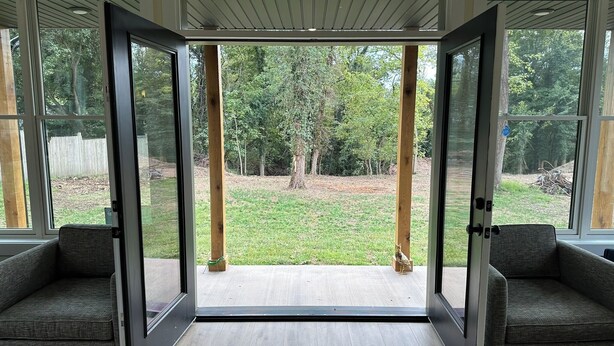 Une pièce dotée d’une grande porte-fenêtre coulissante qui donne sur un jardin avec une pelouse verdoyante et des arbres. On y trouve deux fauteuils gris, recouverts de tissu.