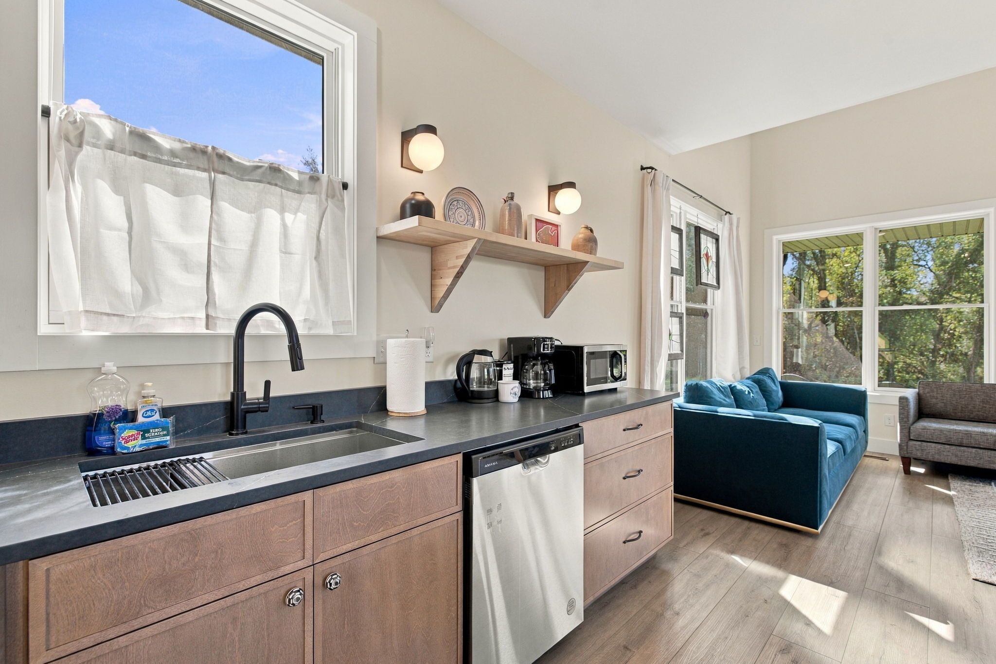 A modern kitchen with a dishwasher, a coffee maker, and a microwave. There is a window with a curtain, and a sofa is visible in the adjacent living area.