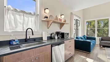 A modern kitchen with a dishwasher, a coffee maker, and a microwave. There is a window with a curtain, and a sofa is visible in the adjacent living area.