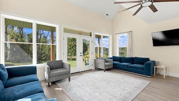 A spacious living room with a large window, a ceiling fan, a flat-screen TV, and a blue sofa.