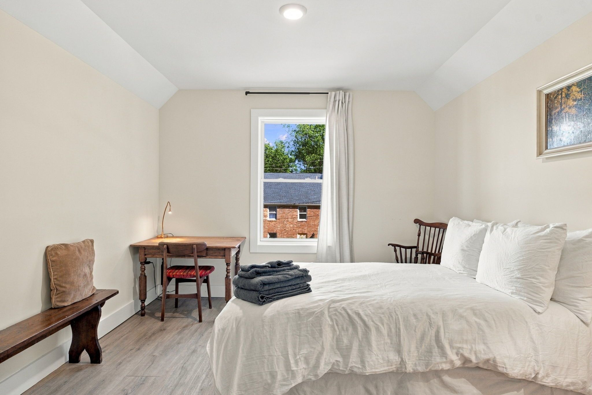 A bedroom with a bed, a desk, a chair, a window, and a painting on the wall.
