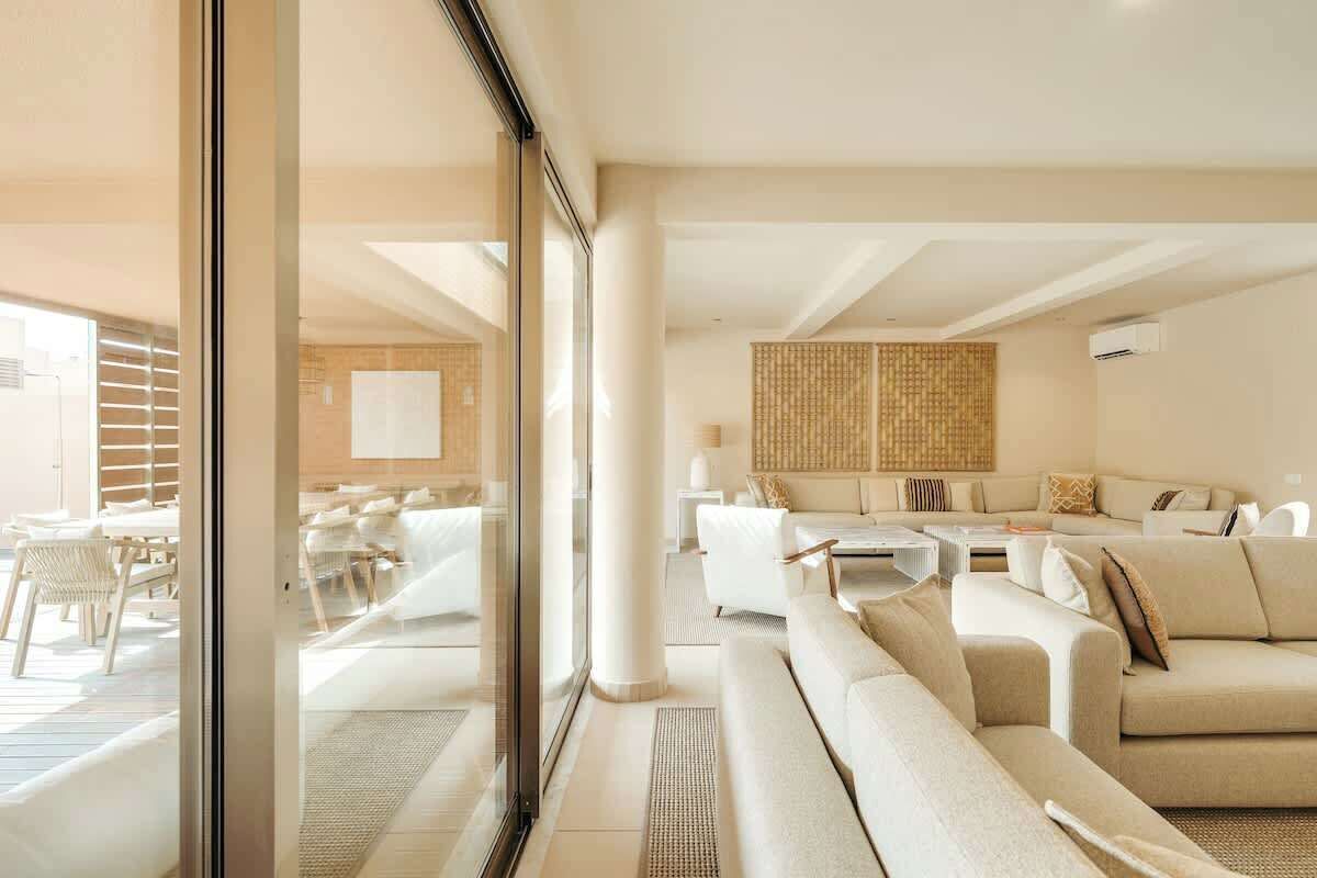 A modern hotel room with a large glass door leading to a balcony, a seating area with sofas and a coffee table, and a large woven wall decoration.