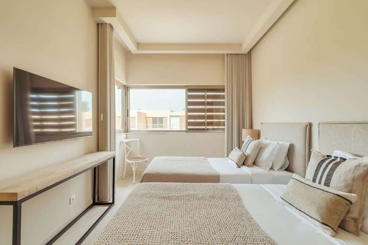 Et hotelværelse med to senge, et fjernsyn ophængt på væggen, et skrivebord og et vindue med persienner.