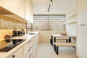 Une cuisine moderne avec un plan de travail noir, des armoires blanches, une fenêtre avec des stores et un petit coin repas comprenant une table et des chaises.