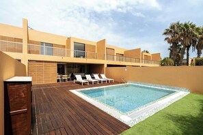 A modern building with a swimming pool, lounge chairs, and palm trees.