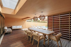 A sunlit outdoor dining area with a wooden table and chairs, a sofa, and a pool in the background.