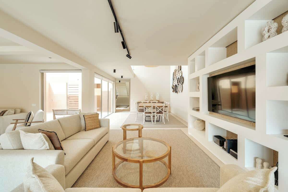 Ein modernes Wohnzimmer mit einer Couch, einem Couchtisch und einem Fernseher. Im Hintergrund befindet sich ein Essbereich mit Tisch und Stühlen.