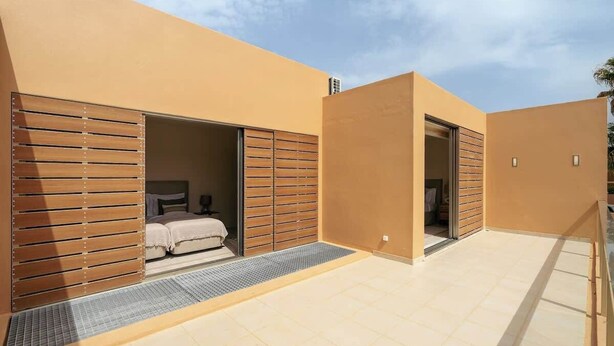 A modern building with a beige exterior, a spacious patio area, and a bedroom with a bed and wooden shutters. - 4BR Villa w/ Private Pool | Salgados | DA’Home (Albufeira)