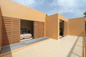 A modern building with a beige exterior, a spacious patio area, and a bedroom with a bed and wooden shutters.