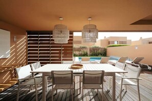 Un vaste espace extérieur aménagé pour les repas, avec une table et des chaises blanches, deux luminaires suspendus et une vue sur une piscine et la verdure en arrière-plan.