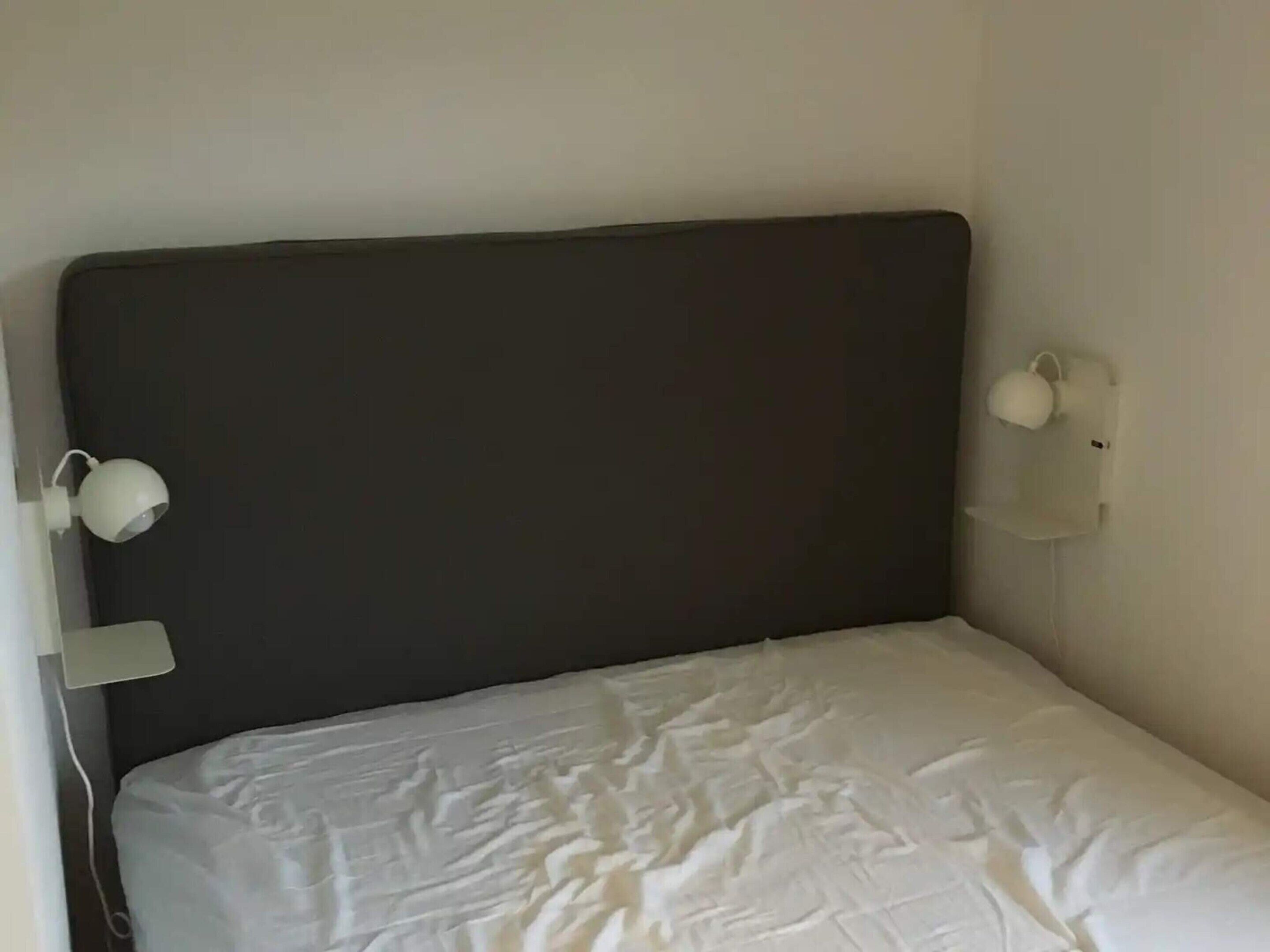 Et lille værelse med en enkeltseng, mørkt sengegavl, hvide sengetæpper og en lampe monteret på væggen.