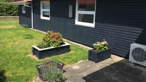 Una casa blu scuro con tetto rosso, due finestre e un piccolo giardino con fiori e una pianta di lavanda.