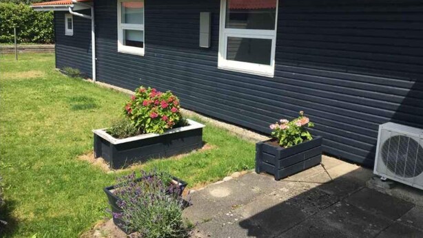 Una casa azul oscuro con techo rojo, dos ventanas y un pequeño jardín con flores y arbustos.