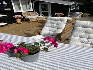 Um pátio com mesa listrada, duas cadeiras brancas com almofadas e um vaso com plantas de flores cor-de-rosa.