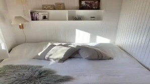 A neatly made bed with pillows and a fluffy rug, a shelf with books and decorative items, and a lamp on the wall. - 5 Star Holiday Home in Dronningmolle (Dronningmolle)