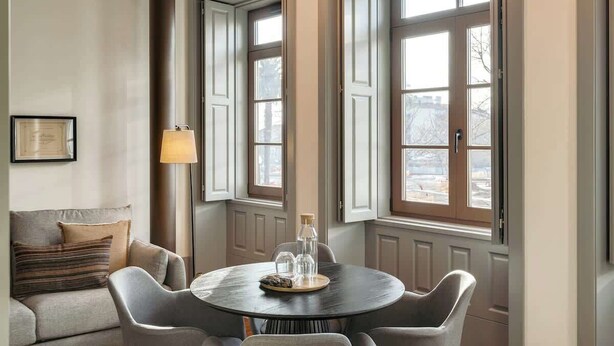 A dining area with a round table, chairs, and a lamp. There are large windows with shutters, a framed certificate on the wall, and a sofa with cushions.