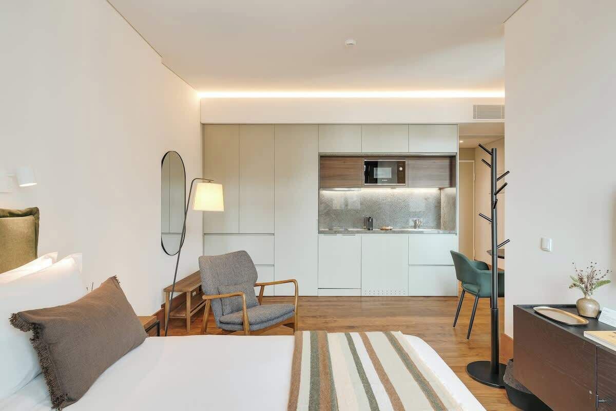 Una moderna camera d'albergo con un letto, una sedia, una scrivania, una lampada e una piccola cucina sullo sfondo.