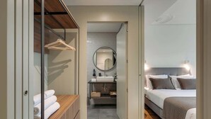 Een moderne hotelkamer met een houten vloer, een groot bed met een geruit plaid, een nachtkastje, een spiegel en een kledingkast met hangende kleding.