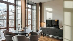 Een moderne woonkamer met een ronde eettafel, grijze stoelen, een donker houten kast, een flatscreen televisie en grote ramen met uitzicht op de stad.