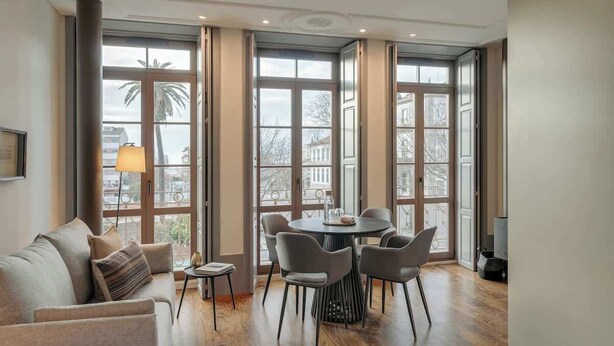 Een moderne woonkamer met een bank, een eettafel met stoelen en grote ramen met gordijnen.