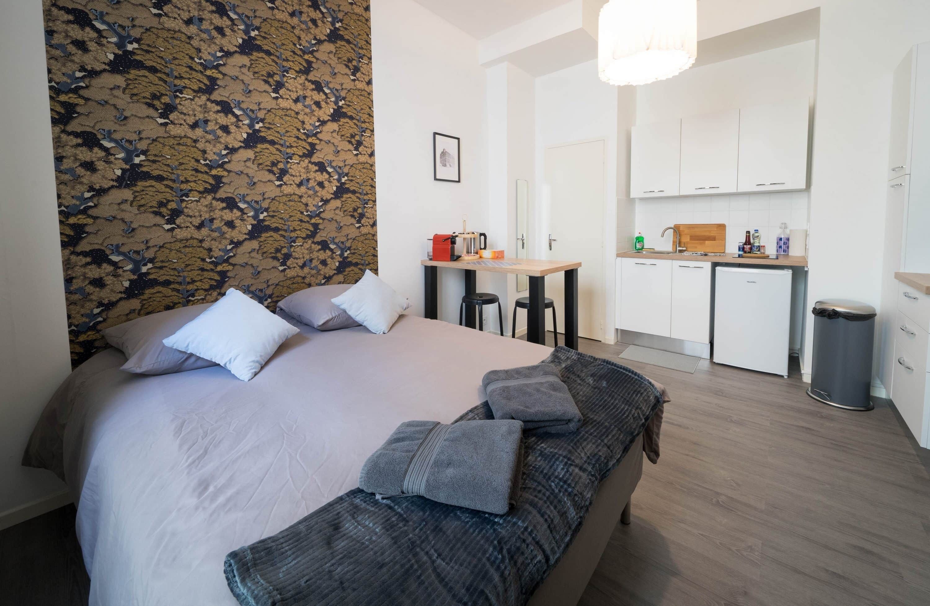 A bedroom with a bed, pillows, a bench, a small table with a kettle, and a kitchenette with cabinets and a trash bin.