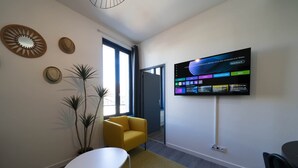A modern room with a flat-screen TV mounted on the wall, a yellow armchair, a small white table, and a potted plant. - Résidence Bardiaux - Chez Mélèze (Vichy)