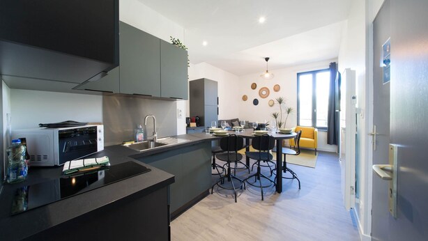 A modern kitchen with a built-in oven, a dining area with a table and chairs, and a bright living space with a sofa and a large window. - Résidence Bardiaux - Chez M élèze (Vichy)