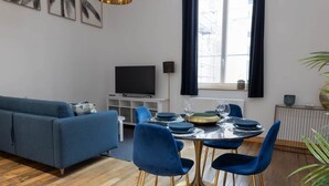 Un salon moderne avec un canapé bleu, une table à manger avec des chaises bleues, un téléviseur à écran plat et une grande fenêtre avec des rideaux.