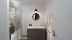 Une salle de bain moderne avec une douche à parois de verre, un miroir rond à cadre noir, un lavabo blanc avec deux tiroirs et un porte-serviettes fixé au mur.