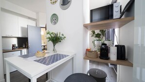 A modern kitchen with white cabinets, a white countertop, and a dining area with a white table and a gray chair. There are decorative plates and a vase with flowers on the wall.