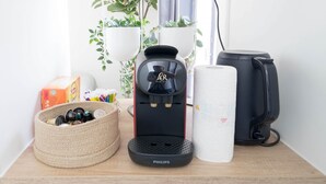 A coffee maker, a basket of toiletries, a paper towel roll, and a black bag on a wooden surface.
