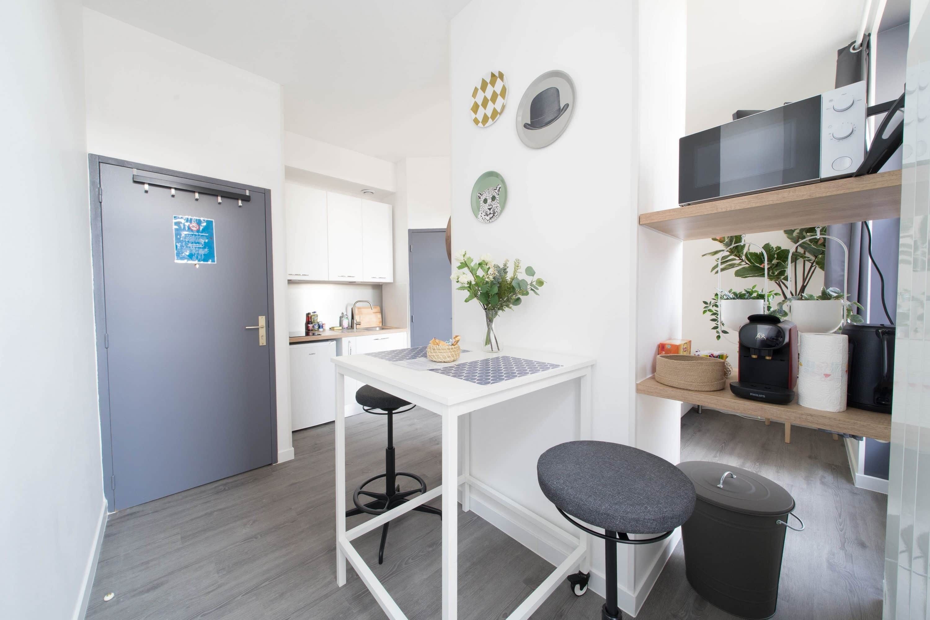 A compact kitchen with a white table, two stools, a small trash bin, and a microwave on a shelf.