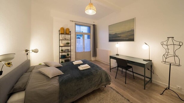 A bedroom with a bed, bedside table, desk, chair, wardrobe, and a window. - Résidence Carnot - Chez Klein (Vichy)