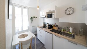 A compact kitchen with white cabinetry, a microwave, a sink, and a clock on the wall. - Résidence Carnot - Chez Majorelle (Vichy)