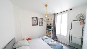 A modern bedroom with a bed, a standing fan, a shelf with towels, and a window with curtains. - Résidence Carnot - Chez Majorelle (Vichy)